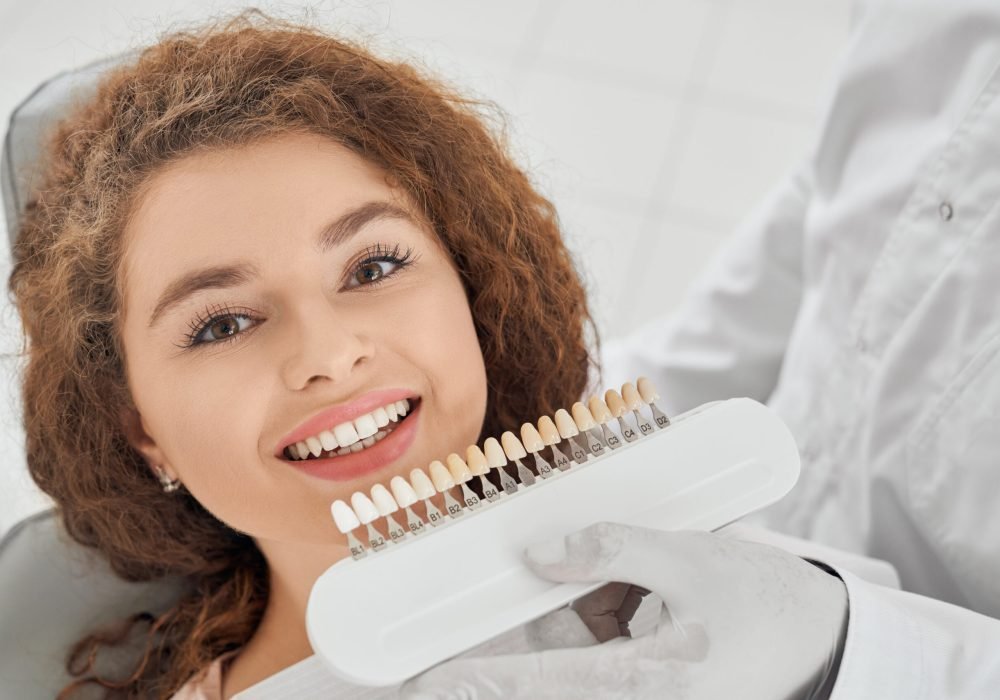 Closeup of beautiful young woman lying on dental chair, looking at camera and smiling while male dentist keeping in hands teeth color range. Girl doing whitening procedure in dental office.